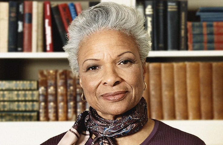 An older Black woman smiles slightly while posing in front of a shelf of books